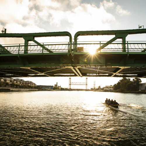 A crew of rowers in a boat on a river in the early morning sunlight. Bridge in the foreground. From the feature doc A Most Beautiful Thing.
