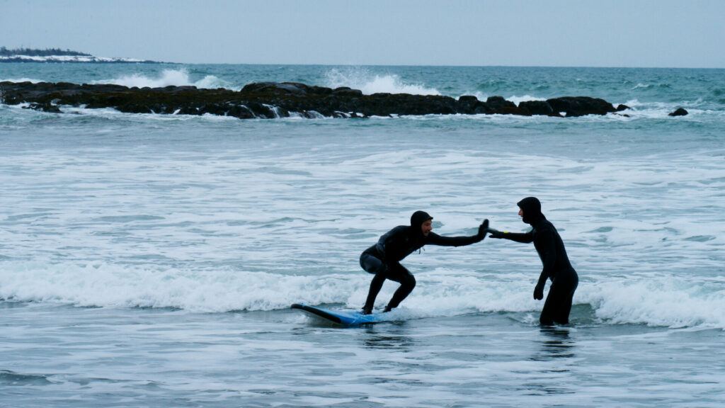 Image of two men in wetsuits high-fiving in the waves. From the short doc 'To Play' by Maika Hearson at CSFF 2024.