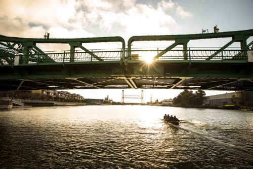 A crew of rowers in a boat on a river in the early morning sunlight. Bridge in the foreground. From the feature doc A Most Beautiful Thing.